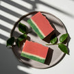 Two handmade watermelon-themed soap bars on a plate with green leaves, casting shadows on a light background.
