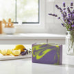 A rectangular bar of lavender and yellow marbled soap sits on a white countertop in a bright kitchen setting, with lemon slices, lavender sprigs, and a vase of purple lavender flowers nearby.