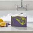 A rectangular bar of purple soap with bright yellow swirls sits on a white bathroom counter in front of a blurred sink and faucet. Lemon wedges and a sprig of lavender are placed beside it.