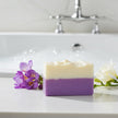 Two-tone Freesia soap bar with purple and white on a bathroom counter with flowers and bathtub faucet in the background.