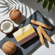 A rectangular layered coconut sandalwood soap bar in off-white, yellow, and brown sits on a dark slate tray, styled with two whole coconuts, a halved coconut, and three wooden pieces of sandalwood on a light marble surface with leaf shadows.