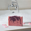 A rectangular bar of pink Dewy Rose  soap with dark purple swirls and fine glitter stands on a white marble surface in front of a blurred bathroom sink and faucet, with scattered pink petals around it.